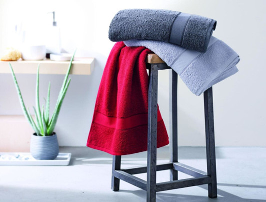 A bath towel display featuring a red, grey, and light grey towel on a black wooden stool, with plants in the background.