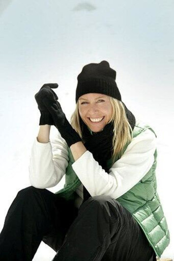 A woman wearing a black beanie and gloves, seated on snow, dressed in a green vest and black pants.