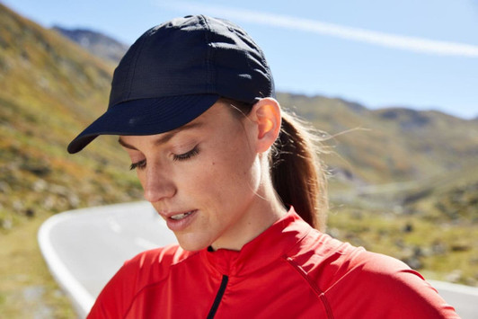 A navy 6 Panel Coolmax® Cap worn by a woman in a red top, with a blurred mountain road in the background.
