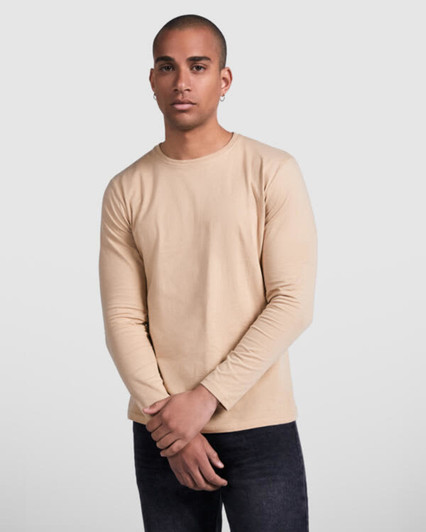 A man wearing a beige long-sleeved t-shirt, standing with arms crossed, against a neutral background.