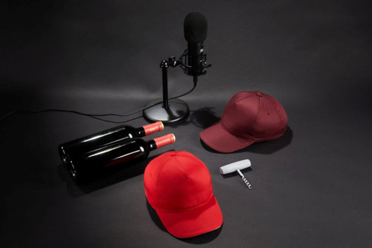 A red cap and a maroon cap are displayed alongside two wine bottles and a microphone, set against a dark background.