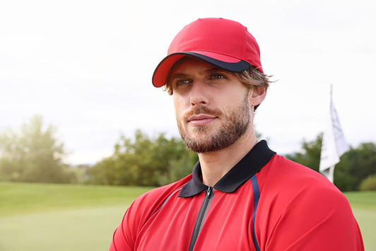 A red 5 panel sports cap positioned on a model outdoors near a golf course. The cap features a logo on the front.