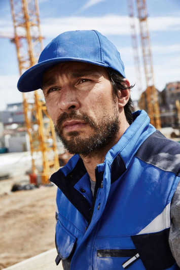 A blue 6 Panel Workwear Cap worn by a man in a blue jacket, set against a construction site background.