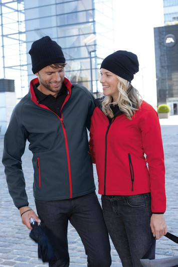 A couple outdoors, wearing a grey jacket with red accents and a black beanie, alongside a red jacket and black beanie.
