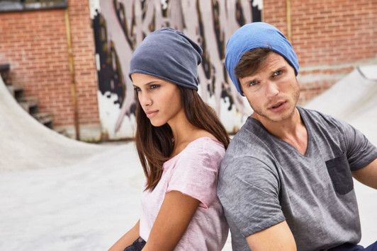A grey and blue beanie is displayed on a male and female model in a casual setting. The background features textured walls and skate ramps.