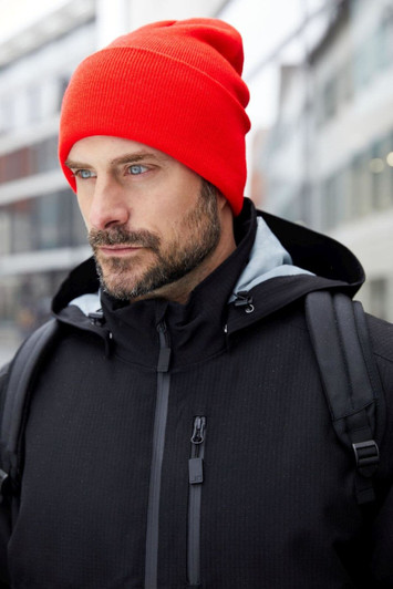 A red high brim beanie worn by a man in a black jacket with a backpack in an outdoor setting.