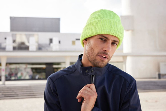A reflective winter beanie in bright neon yellow, worn by a man in a navy jacket, set against a modern architectural background.