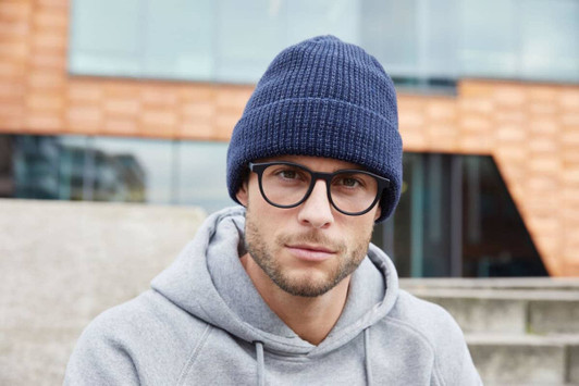 A reflective winter beanie in navy blue, worn by a man in glasses and a grey hoodie, sitting outdoors.