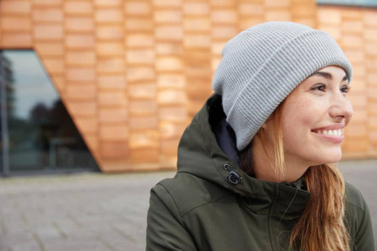 A soft grey knitted winter beanie worn by a woman with long hair, dressed in a green jacket.