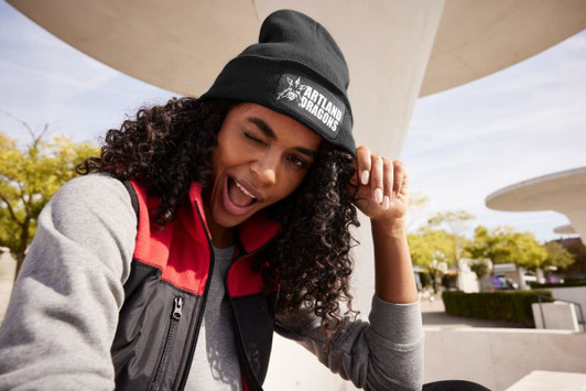 A black knitted beanie with a patch is worn by a person with curly hair, smiling and winking outdoors.