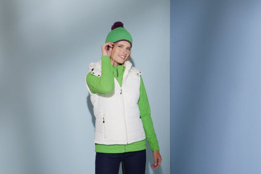 A woman wearing a white vest and a green Pompon Hat with Contrast Stripe, standing against a blue and grey background.