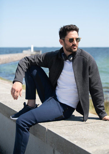 A men's polar fleece jacket in dark grey, worn by a man sitting by the sea, paired with navy pants and sunglasses.