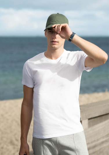A white men's T-shirt displayed outdoors, with a model wearing a green cap and grey shorts by the beach.