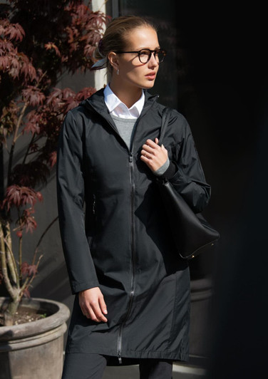 A black rain jacket with a hood, worn by a woman holding a black bag, against a backdrop of plants.