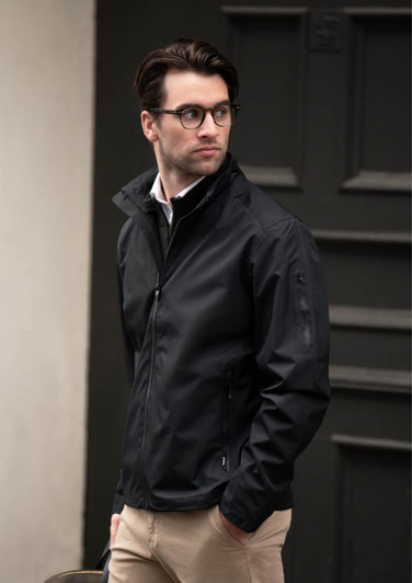 A man wearing a black jacket stands outdoors, paired with light trousers and glasses, against a dark background.