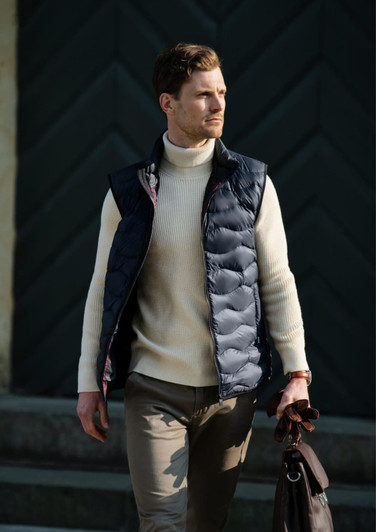 A man wearing a navy quilted vest over a cream turtleneck, holding a brown bag, against a dark green background.
