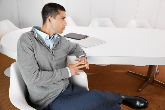 A grey men's zip cardigan worn by a seated man, styled with a blue shirt and sitting at a white table.