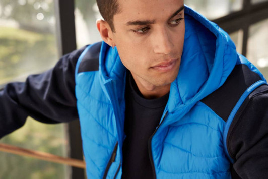 A men's hybrid vest in bright blue with a hood, featuring a quilted design and a navy base layer.