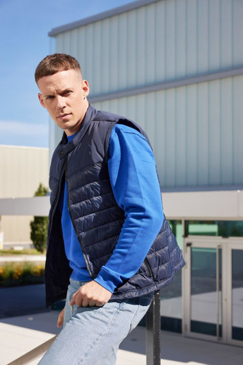 A man wearing a navy padded vest over a blue long-sleeve shirt, standing outdoors near a building. The vest features a logo.