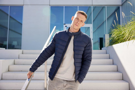 A navy padded jacket worn by a man standing on outdoor steps with modern architecture in the background.