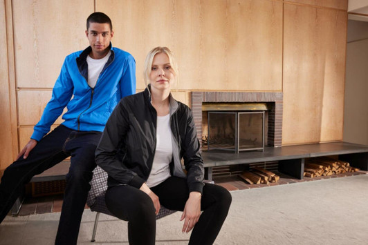 A blue men's blouson jacket and a black women's blouson jacket displayed on models indoors beside a fireplace.