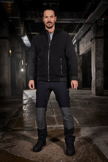 A man stands in a dimly lit industrial space, wearing black and grey workwear pants with a zippered jacket.