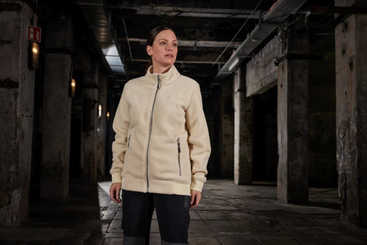 A cream Sherpa jacket with a full zip and side pockets is displayed in an industrial setting.