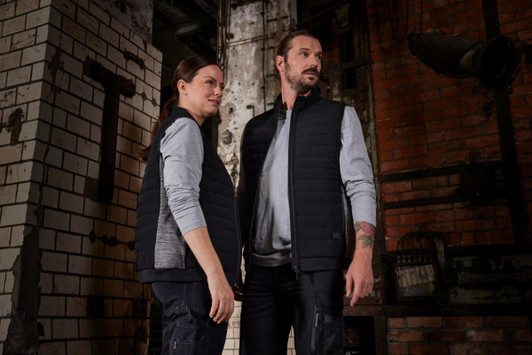 A padded hybrid vest in black, worn by a man and a woman, featuring a logo, set against an industrial backdrop.