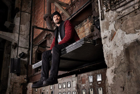 A man wearing a red and black padded hybrid jacket sits on a metal platform in an industrial setting.