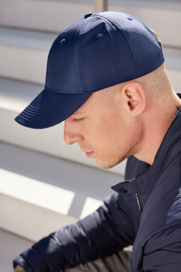 A navy blue 6 panel cap is worn by a man with a shaved head, sitting on stairs. The cap features a logo.