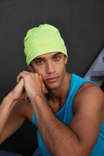 A running beanie in neon yellow worn by a model in a blue tank top, seated against a dark background.