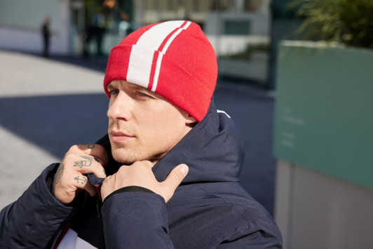 A red winter beanie with white stripes, worn by a person in a dark jacket, set against an outdoor background.
