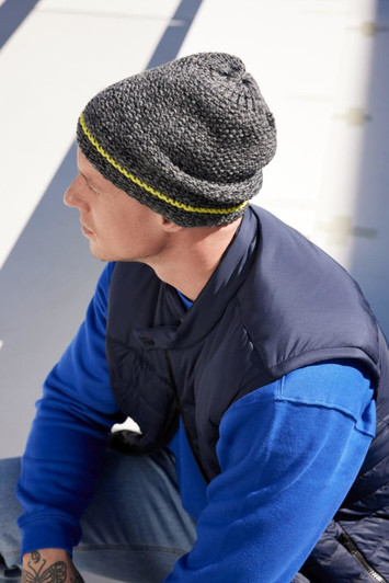 A coarse knitted beanie in grey with a yellow band, worn by a person in a blue sweater and a navy vest.