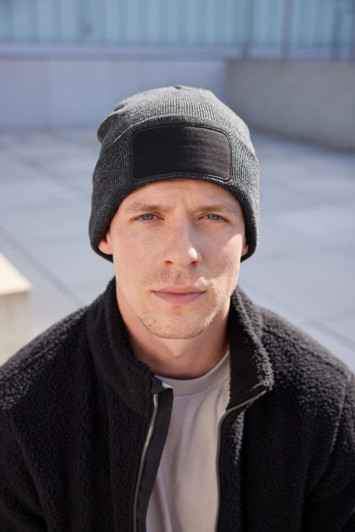 A man wearing a black beanie with a patch, paired with a dark fleece jacket, sits outdoors.