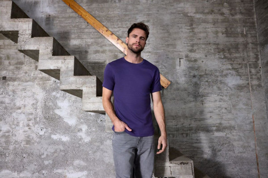 A men's purple T-shirt is displayed on a model standing next to a concrete staircase in a rustic setting.