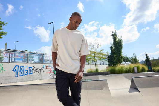 An oversized cream T-shirt worn by a model, set against a sunny outdoor skatepark backdrop.