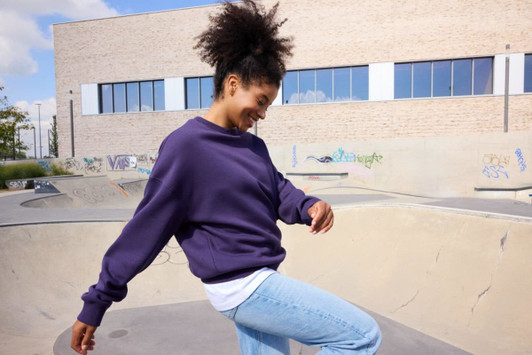 A purple oversized sweatshirt with a relaxed fit, worn by a person smiling near a skate park.