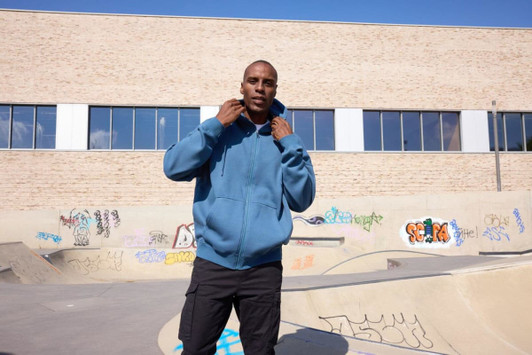 An oversized hooded jacket in blue, worn by a person standing in a skate park with graffiti on the walls.