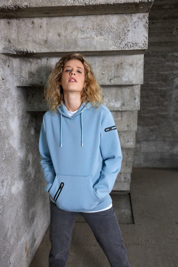 A light blue ladies hoodie with a front pocket, featuring a logo on the sleeve and worn with grey pants.