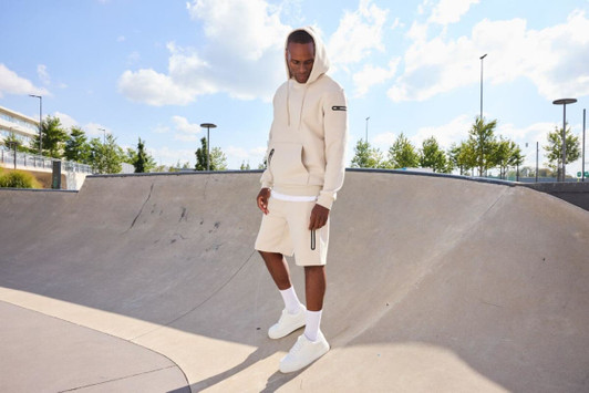An oversized hooded jacket in beige is worn with matching shorts, set in a sunny skatepark. The model stands on a ramp.