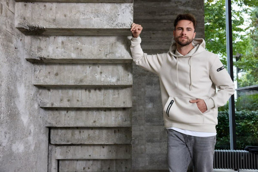 A light beige men's hoodie with a front pocket and a logo, displayed against a modern stone backdrop.