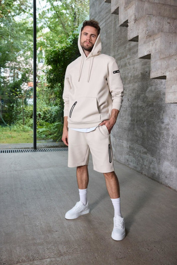 A cream-coloured men's hoodie with a zippered pocket, paired with matching shorts. Features branding on the sleeve.
