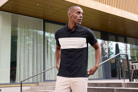 A polo shirt featuring a black base with a cream colour-block design, displayed outdoors. It has a logo.