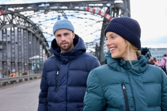 A man in a navy puffer jacket and a woman in a teal puffer jacket, both wearing beanies, stand outdoors.
