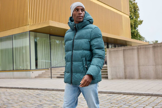 A puffer jacket in teal with a hood, worn by a person outdoors near a modern building.