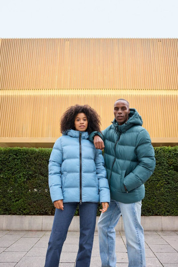 A man in a green puffer jacket and a woman in a blue puffer jacket pose outdoors against a backdrop of greenery.