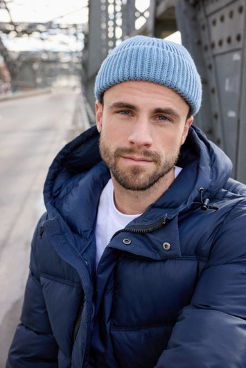 A man wearing a blue fisherman beanie and a navy puffer jacket stands outdoors with a bridge in the background.