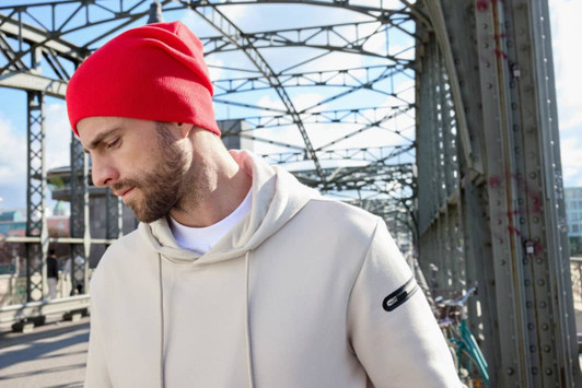 A red casual beanie displayed on a model wearing a beige hoodie, set against an industrial background.
