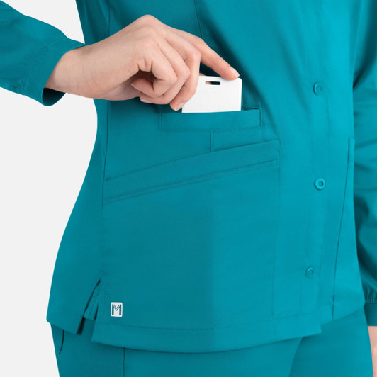 A teal women's round neck snap jacket with a pocket, holding a white card, featuring a logo on the hem.