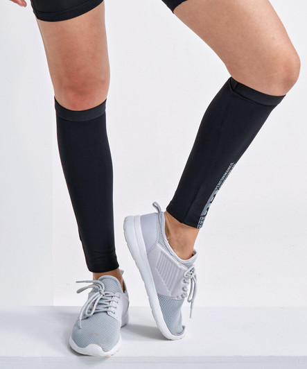 Black compression calf sleeves worn on legs, paired with light grey athletic shoes. The product features a logo.